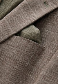 Brauner Blazer mit Struktur und einem gemusterten olivgrünen Einstecktuch. Verfügt über eine dezente Webart und eine Notch-Revers-Detail.