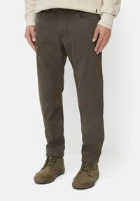 Braune Baumwollhosen mit geradem Schnitt, fünf Taschen und sichtbaren Nähten. Kombiniert mit olivgrünen Stiefeln und einem hellen Sweatshirt.