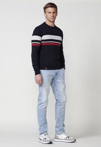 Pulover bleumarin cu dungi albe și roșii, asortat cu blugi albștri deschiși și adidași albi. Puloverul are manșete și tiv ondulate.