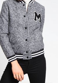 Grå og sort tekstureret varsity-jakke med hvide striber på kraven og manchetterne, der har et sort bogstav "M" patch på fronten.