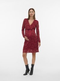 Έγκυος γυναίκα με κόκκινο δαντελένιο φόρεμα μήκους μέχρι το γόνατο με μακριά μανίκια, δεμένη κορδέλα στη μέση, φορώντας μαύρα μποτάκια με τακούνι, στέκεται μπροστά σε λευκό φόντο.