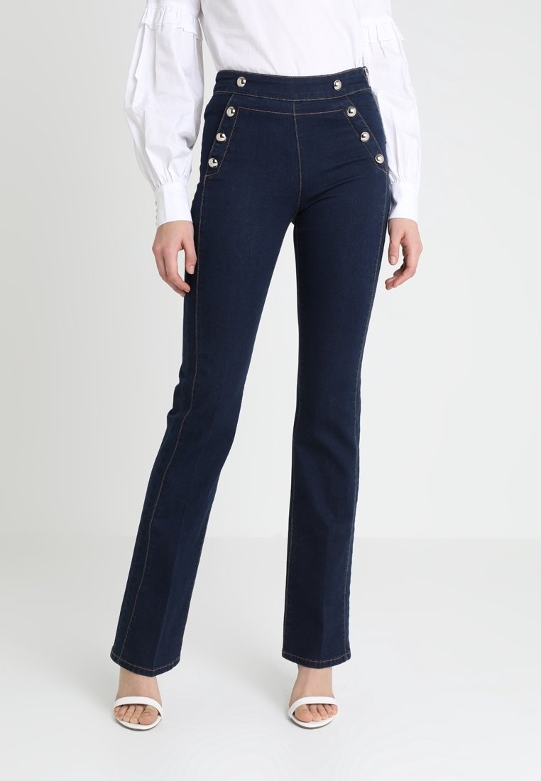 Femme portant un jean bleu foncé taille haute avec des boutons argentés et des talons blancs ouverts à l’avant, associé à un chemisier blanc à manches longues.