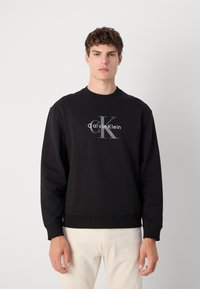 Sweatshirt noir en coton, avec un col rond, des poignets côtelés et un logo graphique à l'avant en blanc et gris.