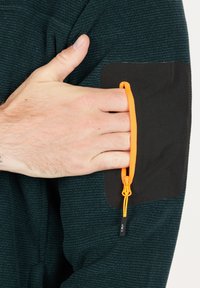 Mörk teal-texturerad tygtröja med en svart lapplag. Har en orange snodd med justerbar snörning för stängning, hand placerad i fickan.