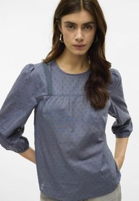 Modrá blůza s tečkami, kulatým výstřihem, nařasenými rukávy, jemnými krajkovými akcenty a strukturovanou tkaninou, s nařaseným předním designem.