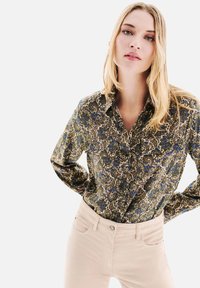 Chemise à boutons florale en motifs vert foncé, bleu et crème ; présente un col et des manches longues ; associée à un pantalon skinny beige clair.