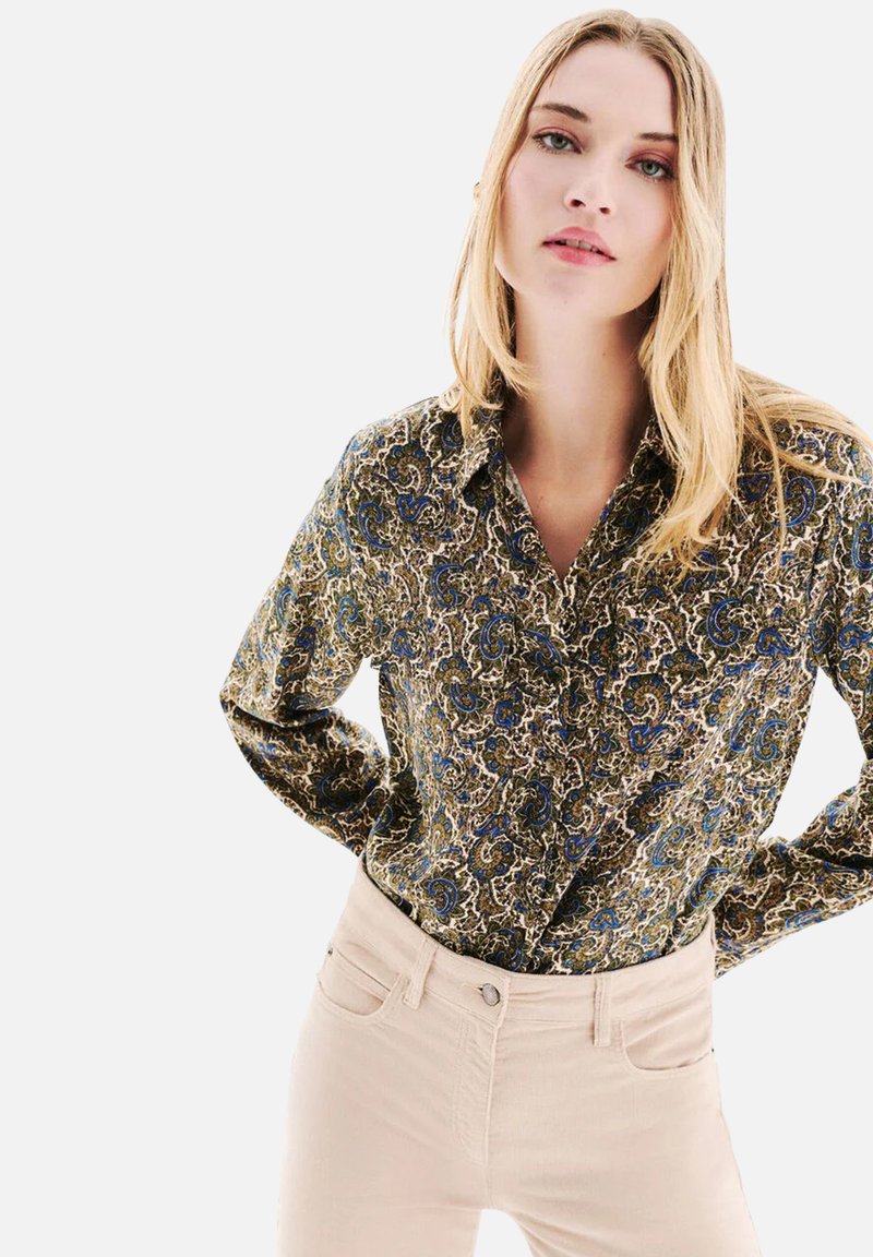 Chemise à boutons florale en motifs vert foncé, bleu et crème ; présente un col et des manches longues ; associée à un pantalon skinny beige clair.