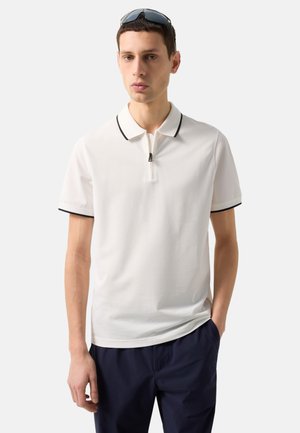 Man draagt een witte korte mouw polo met zwarte bies en rits, donkere broek en zonnebril op zijn hoofd, staand tegen een effen achtergrond.