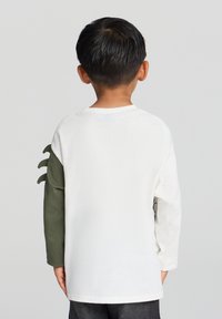 Koszula z długim rękawem ma białą koronkę z zielonym rękawem, teksturowane detale przypominające pazury oraz okrągły dekolt.