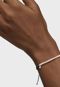 Pulsera de perlas en una muñeca de piel oscura, con pequeñas perlas blancas ensartadas en un cordón verde oscuro y un acento de charm plateado.