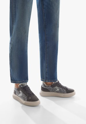 Sneakers grigi con tessuto grigio testurizzato, accenti metallici e suola in gomma. Abbinati a jeans blu, leggermente affusolati alla caviglia.