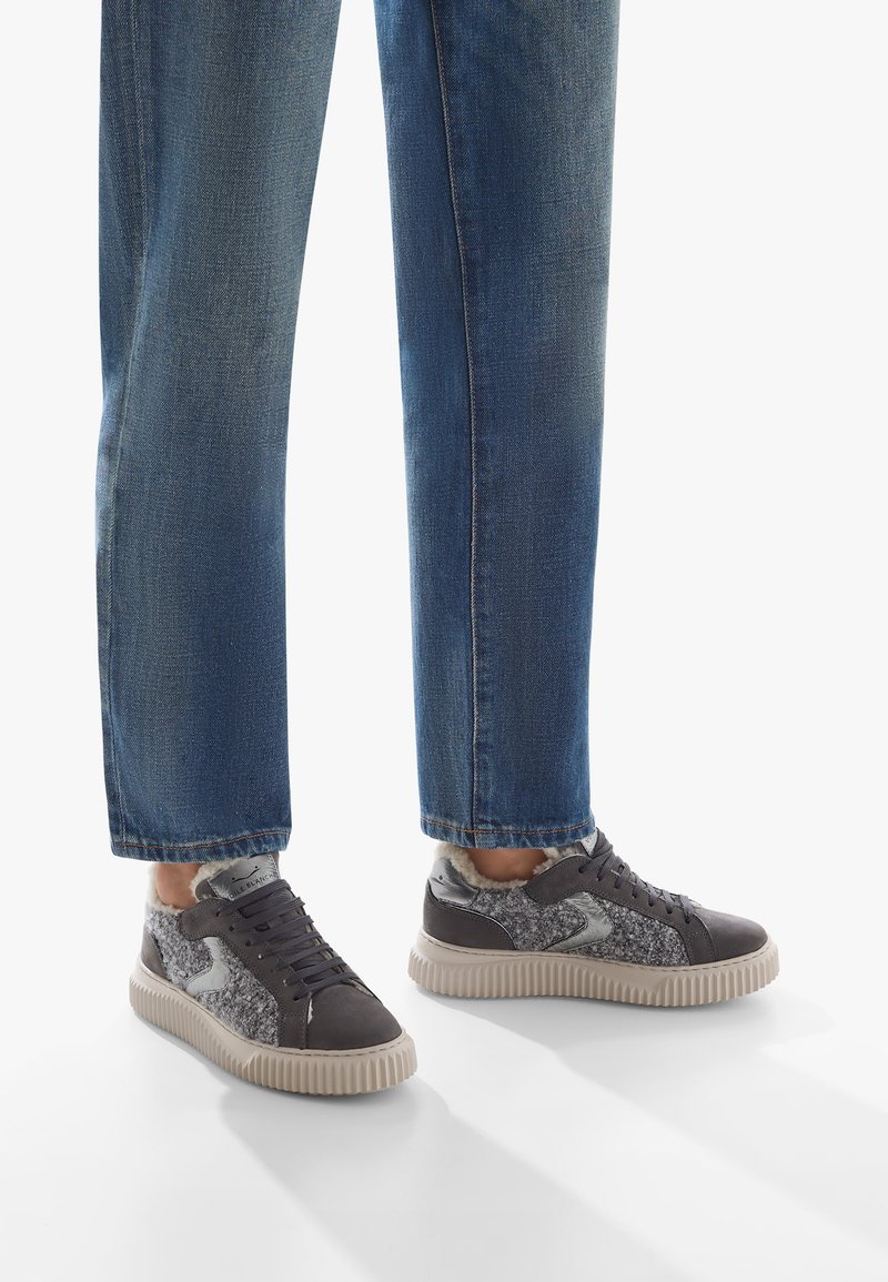 Sneakers grigi con tessuto grigio testurizzato, accenti metallici e suola in gomma. Abbinati a jeans blu, leggermente affusolati alla caviglia.
