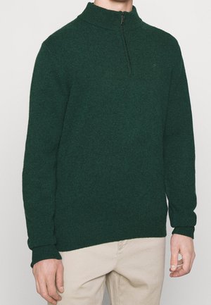 Mann trägt einen dunkelgrünen Strickpullover mit Viertel-Reißverschluss am Kragen und beige Hosen vor einfarbigem Hintergrund.