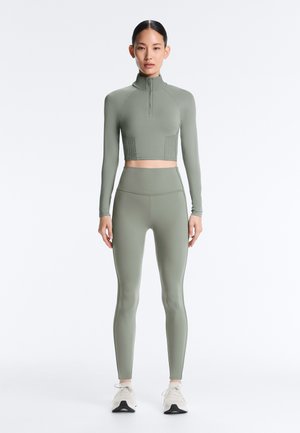 Vrouw die recht vooruit staat, gekleed in een bijpassende olijfgroene cropped top met lange mouwen en ritssluiting, gecombineerd met een high-waist legging en witte sneakers.