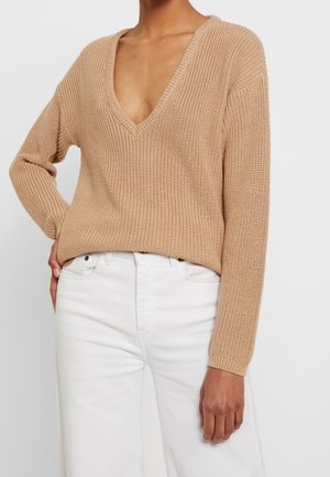 Personne portant un pull beige en maille à col en V et un pantalon blanc taille haute, une main reposant sur la hanche sur un fond uni.