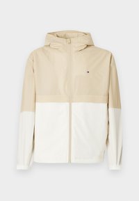 Beżowa i kremowa kurtka przeciwwiatrowa z kapturem, pełnym zamkiem błyskawicznym, elastycznymi mankietami oraz logo na klatce piersiowej. Gładka faktura, lekki materiał, sportowy design.