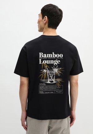 Homme portant un t-shirt noir avec le texte « Bamboo Lounge », un verre à cocktail et un graphisme de plante de bambou au dos sur un fond uni.