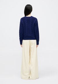 Σκούρο μπλε πλεκτό πουλόβερ με σχέδιο καλωδίου στο πίσω μέρος, συνδυασμένο με κρέμα φαρδιές παντελόνες. Μαλακή υφή και άνετη εφαρμογή.