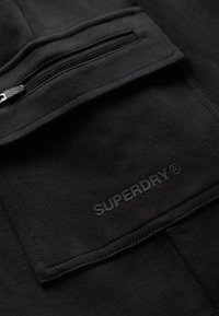 Czarna tkanina z kieszenią na zamek błyskawiczny i wytłoczonym logo "SUPERDRY". Tekstura jest gładka o matowym wykończeniu.