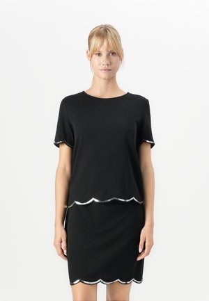 Femme portant une robe noire à manches courtes avec des bordures en forme de coquille argentées aux manches, à l'ourlet et à la taille, se tenant devant un fond blanc.