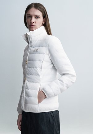 Donna che indossa una giacca trapuntata bianca con collo alto e logo EA7, in piedi con una mano in tasca contro uno sfondo neutro.