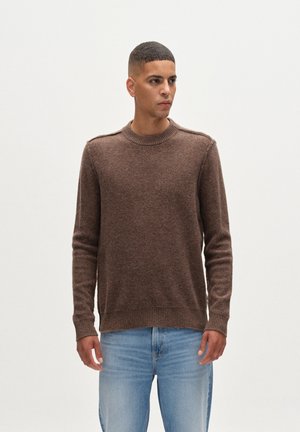 Pulover tricotat, maro, cu gât rotund și manșete cu dungi, asortat cu blugi din denim de un albastru deschis. Design simplu, textură moale și material de greutate medie.