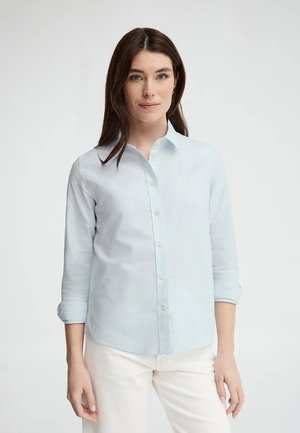 Donna con camicia azzurra a maniche lunghe arrotolate e pantaloni bianchi, in piedi davanti a uno sfondo chiaro e semplice.