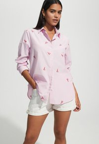 Striped pink button-up shirt featuring red lobster embroidery, long sleeves rolled at the cuffs, paired with white shorts.