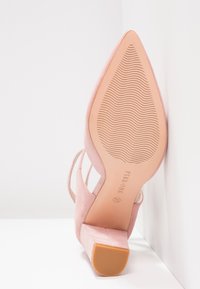 Chaussure à talon haut rose en daim, à bout pointu, positionnée sur le côté, montrant la semelle texturée et le talon carré sur un fond blanc d'angle.