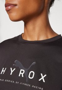 Camiseta deportiva negra con cuello redondo, textura suave y un diseño de logo reflectante que presenta la palabra "HYROX" en blanco y un gráfico de un animal saltando.