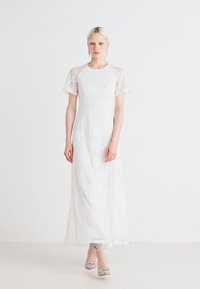 Valkoinen pitsinen maksimekko lyhyillä läpinäkyvillä hihoilla, istuva bodice ja valuva helma. Pehmeä rakenne, jossa on monimutkaisia kukkakuviota. Hopeakengät.