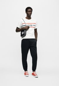 Λευκό μπλουζάκι με κόκκινες και μπλε ρίγες και το κείμενο «CONVERSE ALL STAR», συνδυασμένο με μαύρο παντελόνι και κόκκινα αθλητικά παπούτσια. Μαύρο τσαντάκι χιαστί.