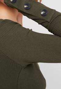 Olivenfärgad ribbad topp med långa ärmar, utrustad med svarta knappar vid ärmslutet och en figurnära design. Texturen är snug och elastisk.