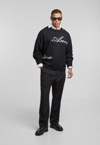 Suéter de punto negro con texto blanco en estilo de escritura, combinado con una camisa blanca de cuello, pantalones negros y zapatos negros con suela gruesa.