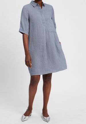 Frau trägt ein locker sitzendes, knielanges, hellblaues Leinenkleid mit hochgekrempelten Ärmeln und silbernen spitzen Ballerinas, steht vor weißem Hintergrund.