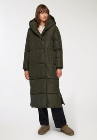 Lungo cappotto imbottito verde oliva con cappuccio, caratterizzato da un design trapuntato, fessure laterali e una cerniera frontale. Indossato sopra jeans scuri e pantofole color sabbia.