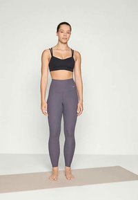 Mustap ultratiimi urheilbra ja harmaat korkeavyötäröiset leggingsit, joilla on sileä pinta ja hienovarainen Nike-logo vyötäröllä. Paljain jaloin joogamatolla.