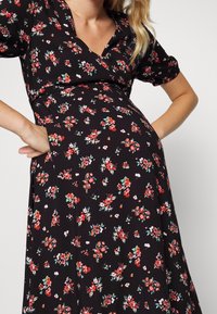 Femme enceinte portant une robe noire avec des motifs floraux rouges et blancs, les mains posées sur les hanches.