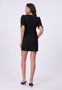Vestido negro corto con mangas abullonadas y detalles de lazo ajustables. Presenta una cintura ceñida y una pequeña abertura en forma de lágrima en la parte de atrás. Textura suave.