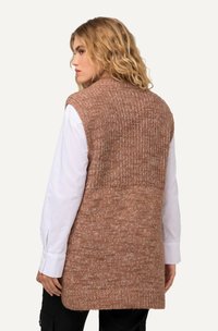 Gilet senza maniche lungo fino al ginocchio in tessuto a maglia marrone testurizzato, con un filo più liscio nella parte superiore della schiena. Indossato sopra una camicia bianca a maniche lunghe.