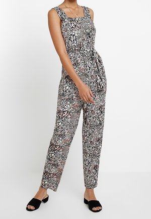 Kvinde iført ærmeløs jumpsuit med dyreprint, bælte i taljen og sorte åbne slidesandaler, stående mod en ensfarvet hvid baggrund.