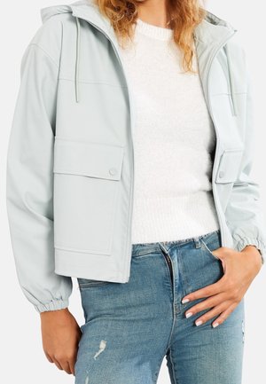 Vrouw draagt een lichtgrijze capuchonjas over een witte trui en blauwe versleten jeans, staat met één hand in haar zak.