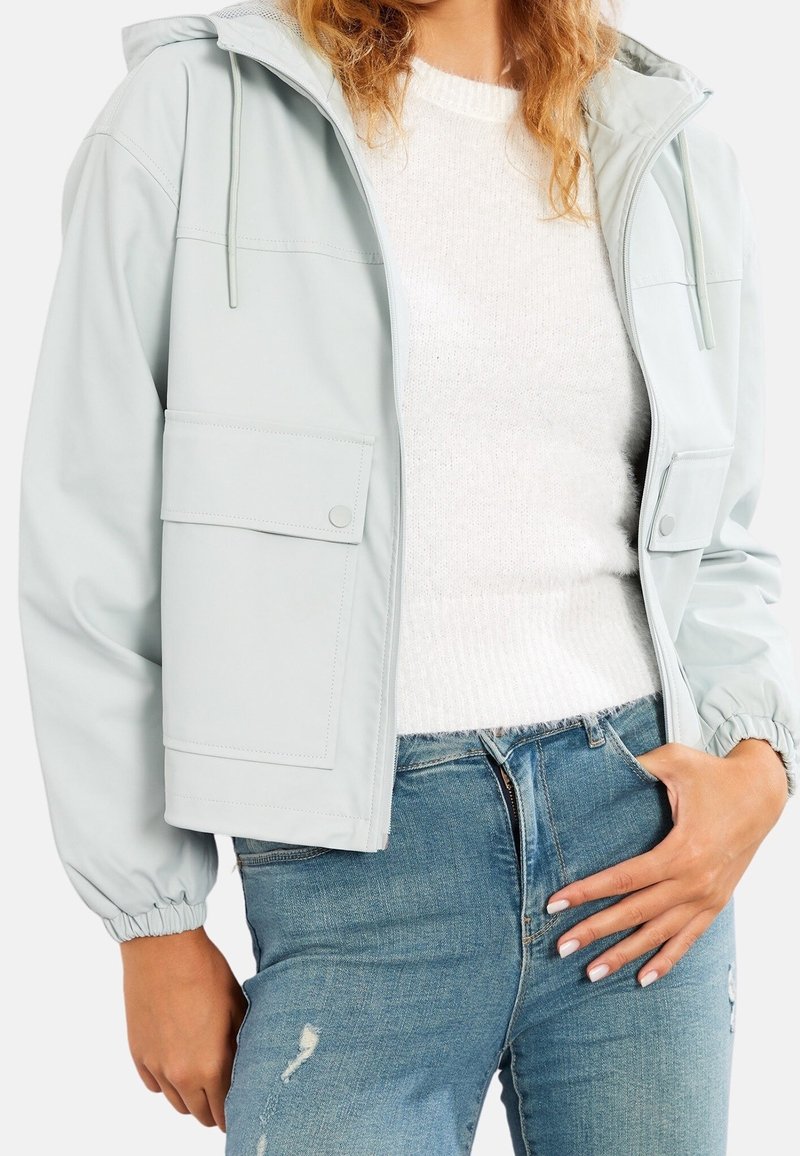 Femme portant une veste à capuche gris clair par-dessus un pull blanc et un jean bleu déchiré, debout avec une main dans la poche.