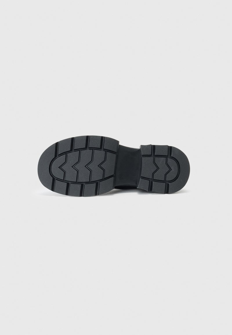 Semelle de chaussure noire avec des motifs de semelle profonds, incluant des chevrons et des rainures rectangulaires, sur un fond gris clair uni.