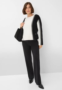 Femme debout portant un pull coloré en noir et blanc, un pantalon noir, des chaussures noires à talons pointus et portant un sac à épaule noir.