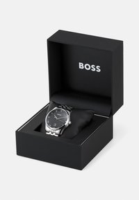 Montre en acier inoxydable argenté avec un cadran noir et des marqueurs en chiffres romains, présentée dans une boîte noire avec le nom de la marque "BOSS" en relief.
