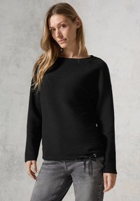 Schwarzer, strukturierter Langarmpullover mit weitem Ausschnitt und Kordelzugdetails am Saum. Getragen mit grauen Jeans.