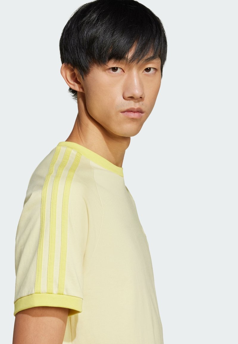 T-shirt gialla a maniche corte con colletto a coste e tre strisce verticali di una tonalità più chiara sulle spalle. Texture del tessuto liscia.