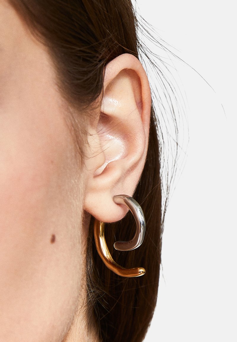 Pendientes de aro de oro y plata con un diseño curvado, que presentan texturas contrastantes, suaves y brillantes, llevados en la oreja.