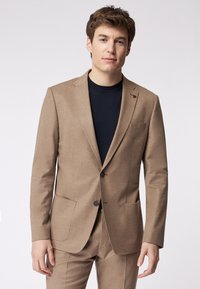 Brauner maßgeschneiderter Blazer aus strukturiertem Stoff, mit Reverskragen, zwei vorderen Taschen und einer einzelnen Knopfschluss. Getragen über einem marineblauen Hemd.
